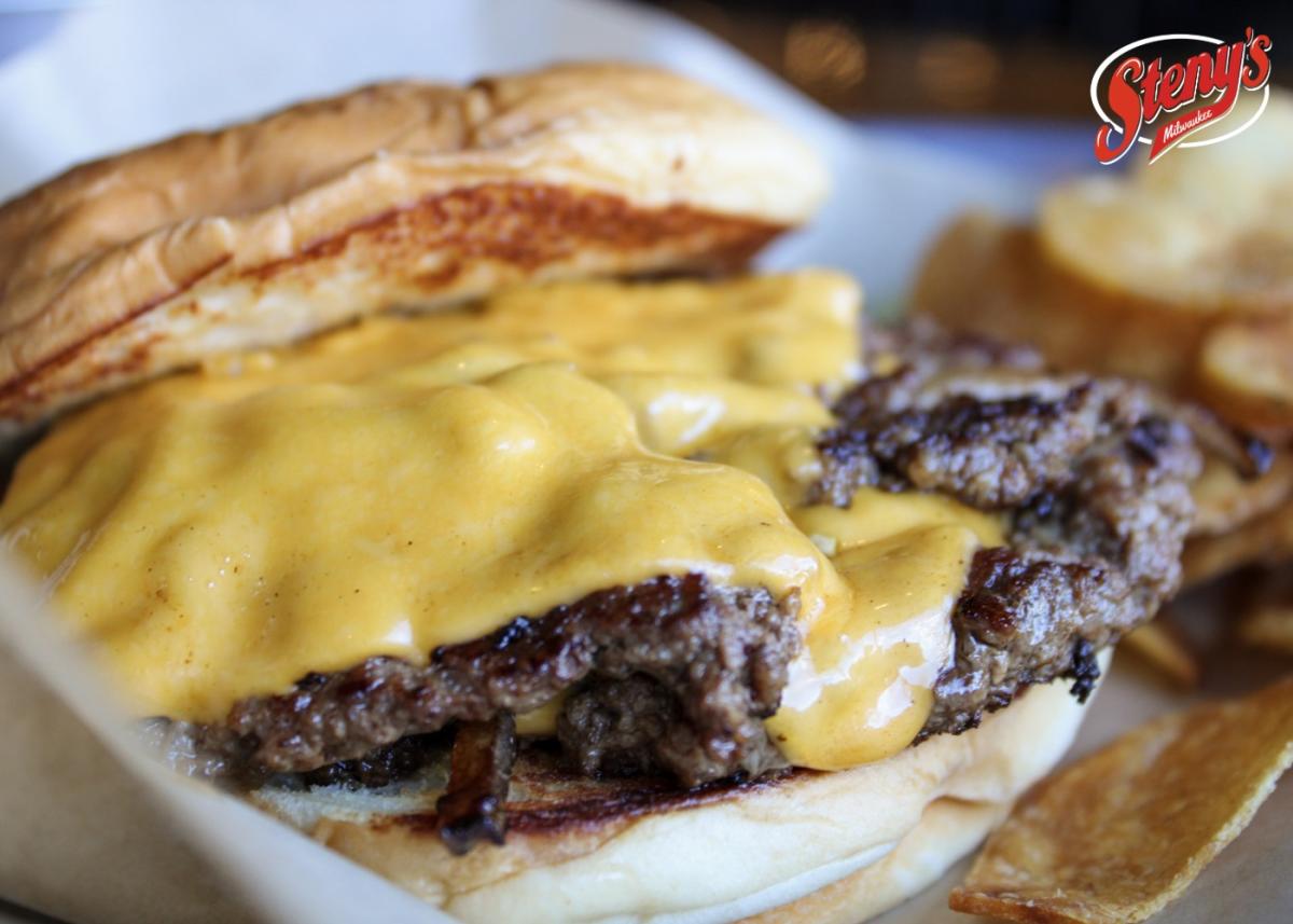 Close up of a double cheeseburger with two beef patties and melted cheese on a toasted bun, served with potato chips on a plate, with the Steny’s Milwaukee logo visible in the corner.