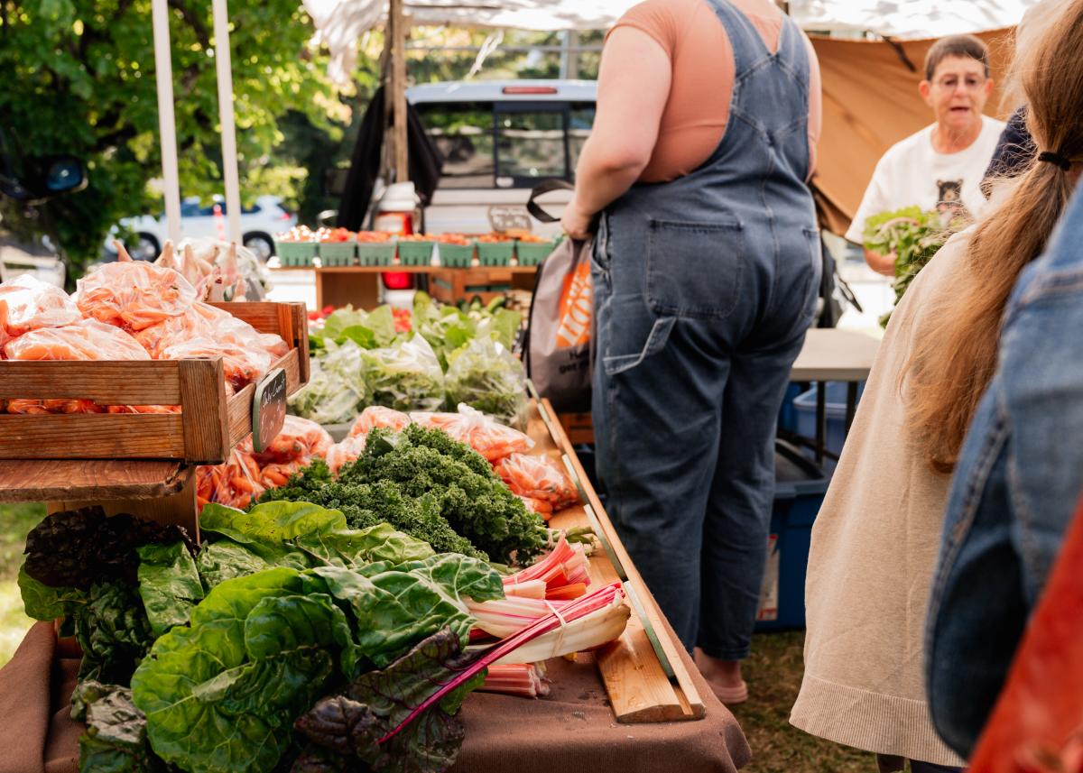Visitors browse a sunny outdoor farmers’ market stall filled with fresh local produce, including leafy greens, carrots, and squash, under white canopy tents.