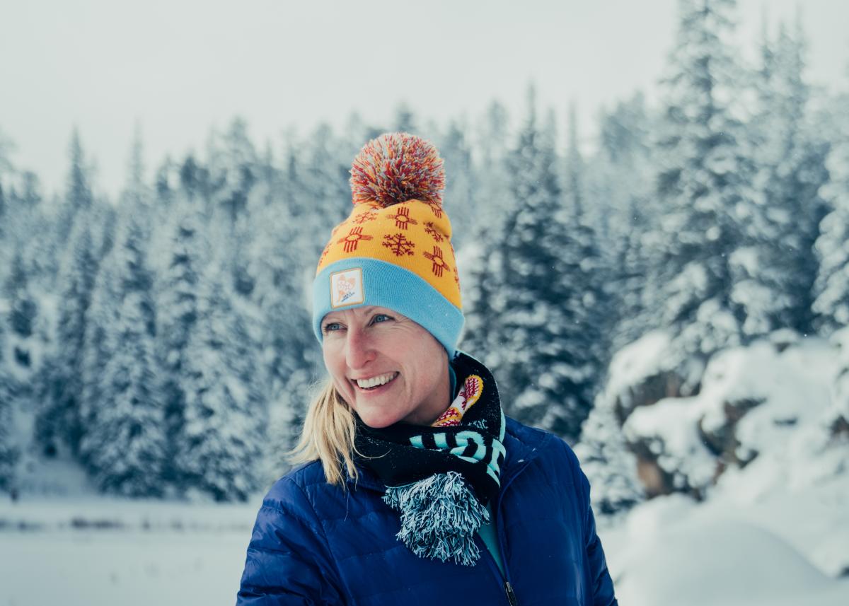 Portrait of Whitney Spivey wearing a yellow and blue NM Zia hat, black scarf and a blue jacket with snow covered trees as a backdrop