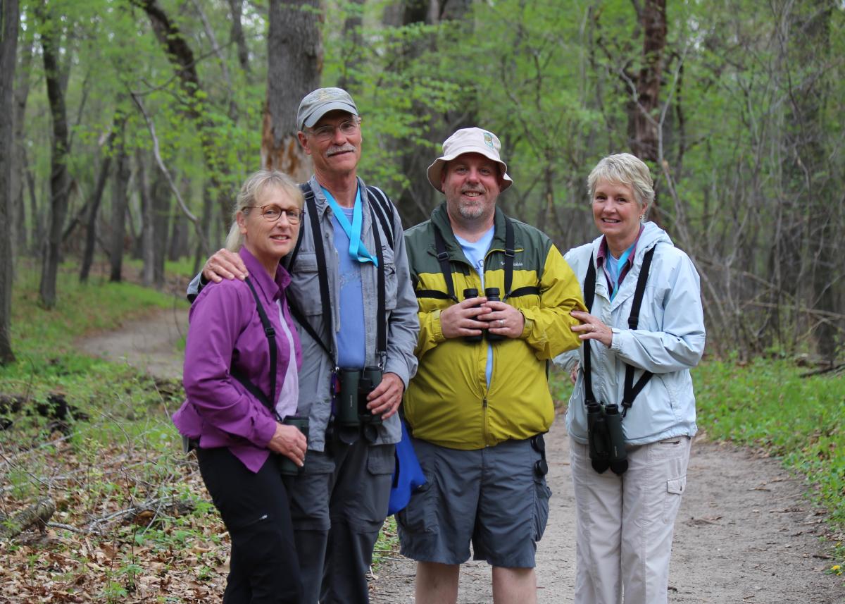 A group of four people stand together on a trail. They are looking at the camera, smiling. Woods are in the background