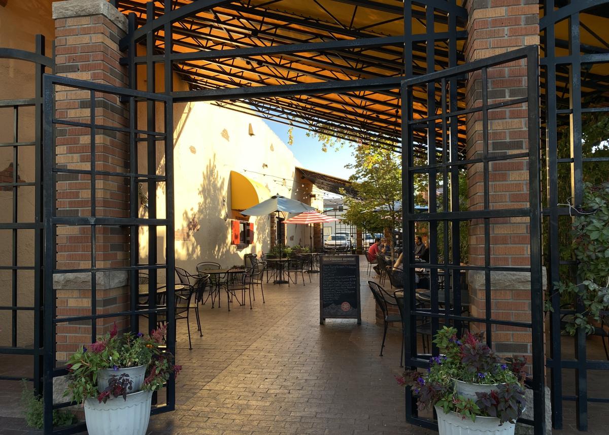 An inviting outdoor patio at Thirsty’s Burgers & Beer with an iron gate entrance, brick columns, yellow awnings, and shaded seating. People sit under umbrellas, creating a relaxed atmosphere.