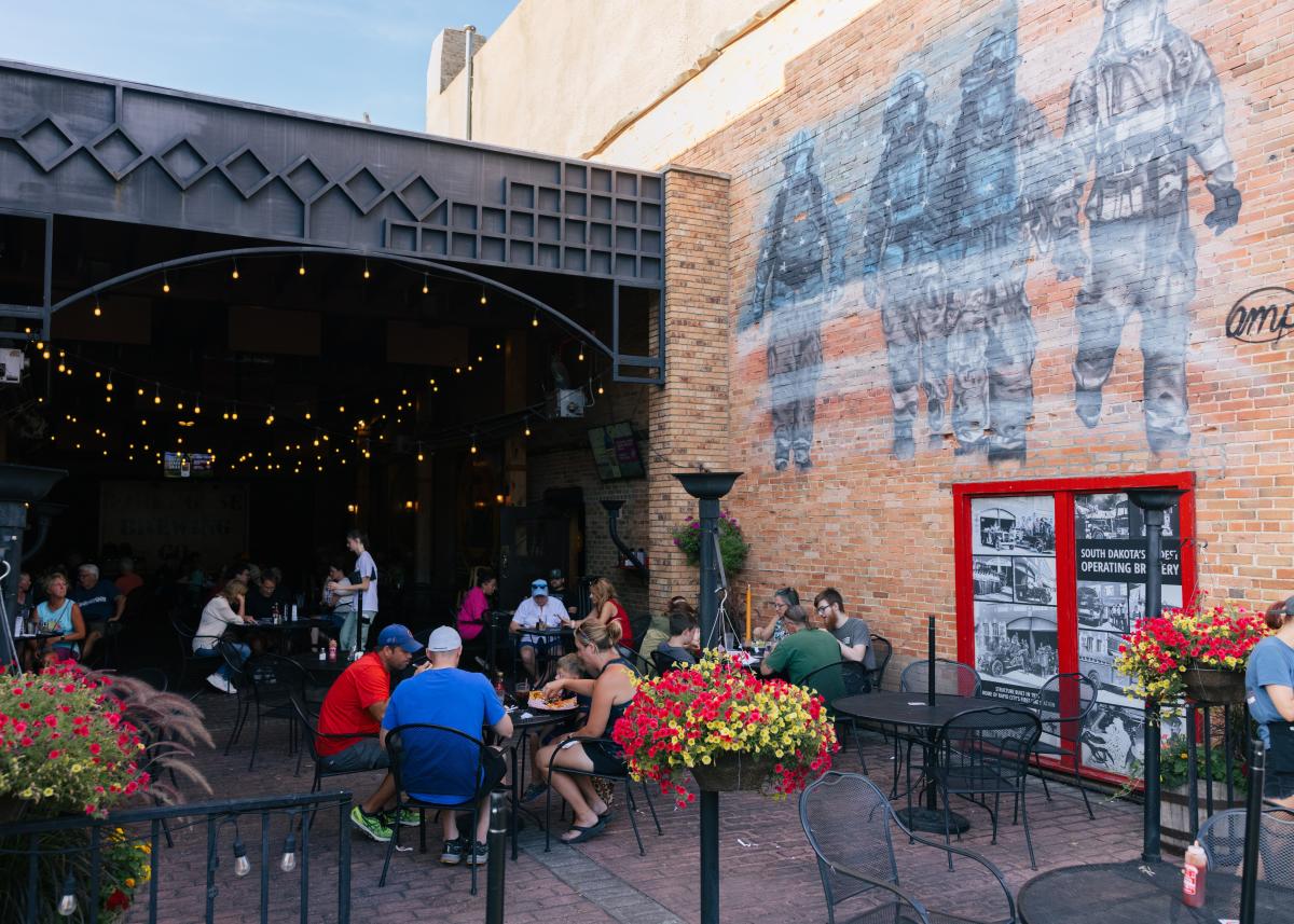Outdoor patio at Firehouse Brewing Co. with people dining under string lights. A large mural of firefighters adorns the brick wall. Hanging flower baskets add color.