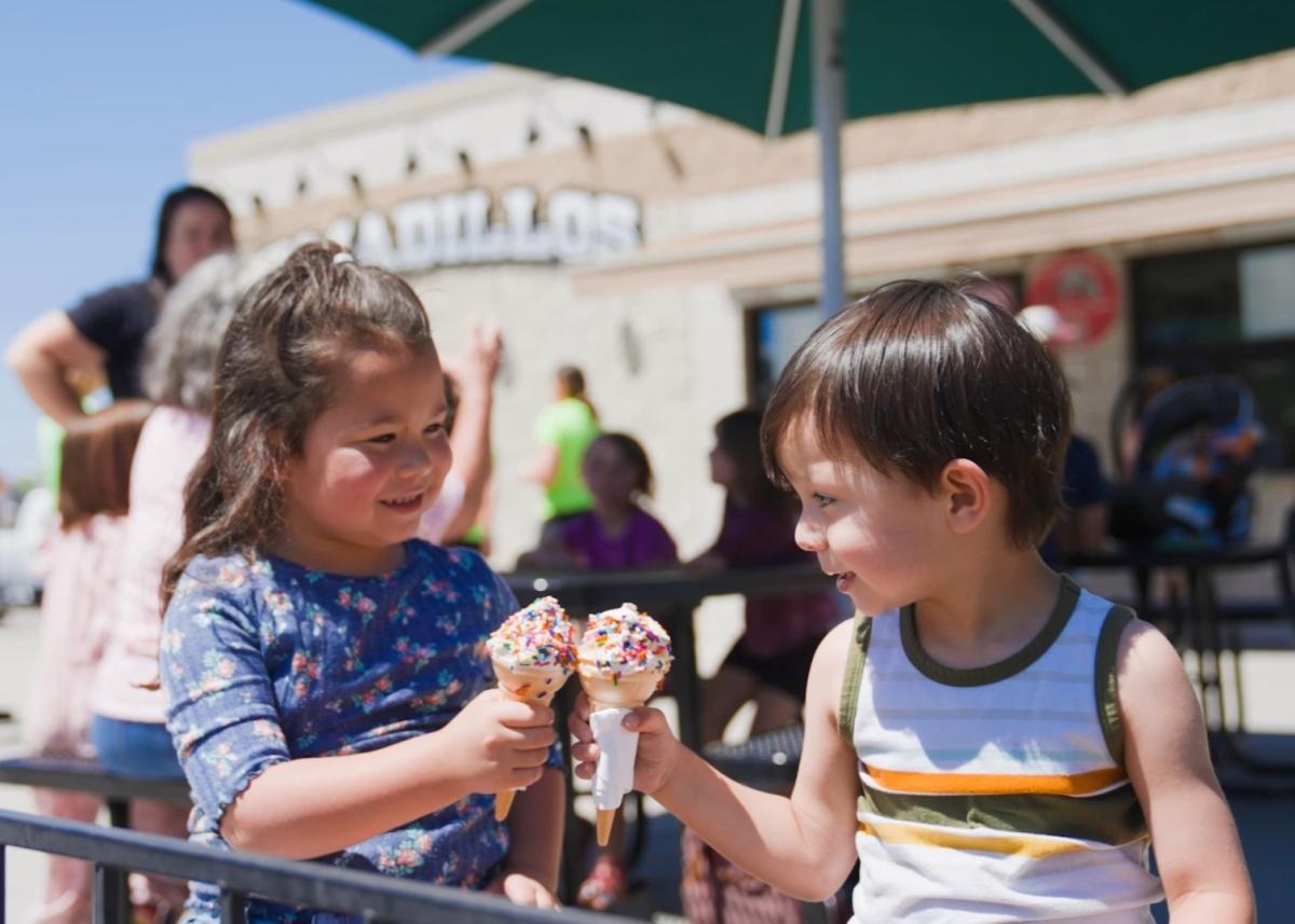 Children smile while toasting ice cream cones with colorful sprinkles. They are outside Armadillos Ice Cream Shoppe with others in the sunny background, creating a joyful atmosphere.