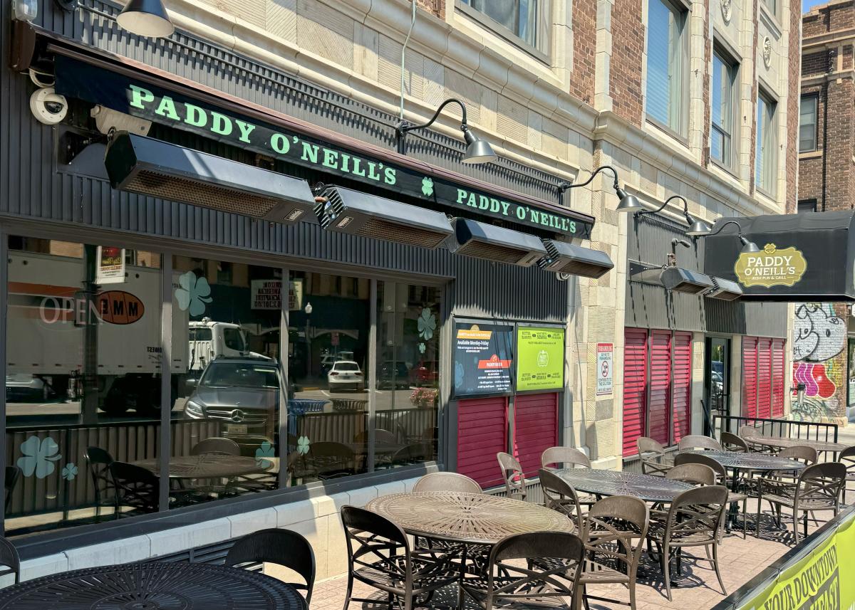 Outdoor seating of Paddy O'Neill's pub with round tables and chairs on a city street. Green signs and umbrellas create a welcoming, lively atmosphere.