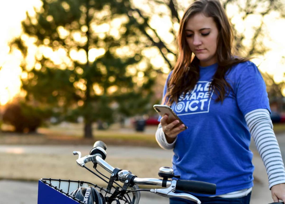 A girl is wearing a blue shirt with a "Bike Share ICT" logo on it standing over a bike