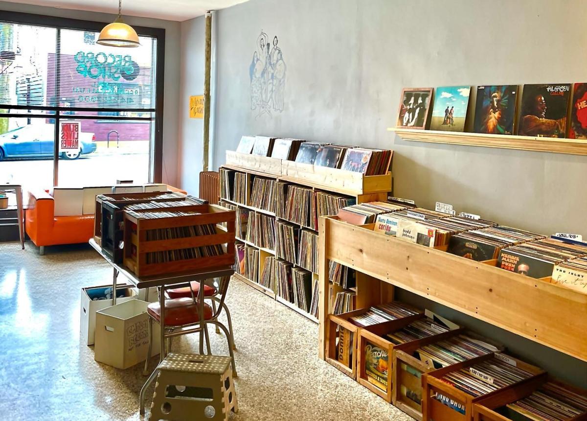A small, laid-back record shop features wooden bins filled with vinyl along the walls and in the center of the room. Sunlight filters through the front window, where a seating area with chairs and a couch sits near the entrance, giving the space a relaxed, neighborhood feel.