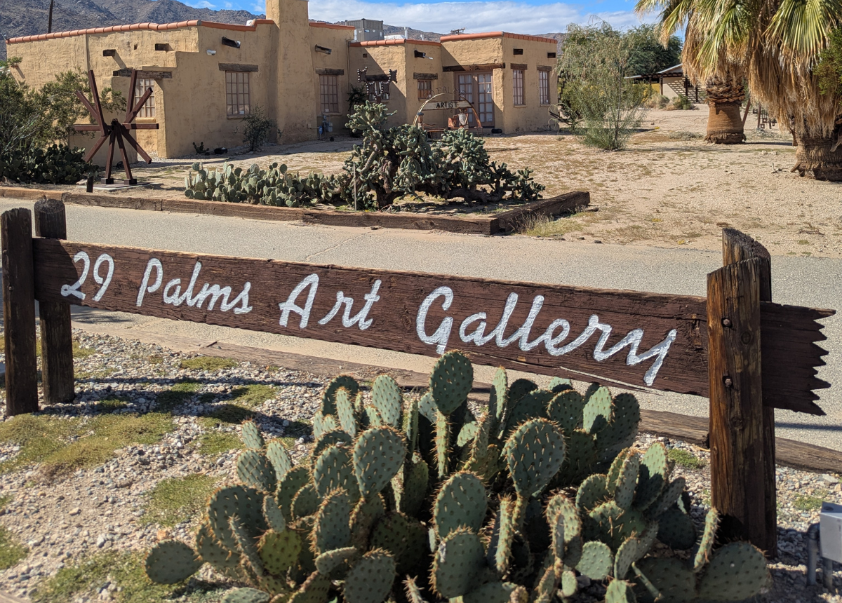 Exterior of the 29 Palms Art Gallery in Twentynine Palms, California, featuring the gallery's wooden entrance sign, desert landscaping, cacti, and adobe-style building, where a Fourth of July celebration with art activities and fireworks viewing will take place.