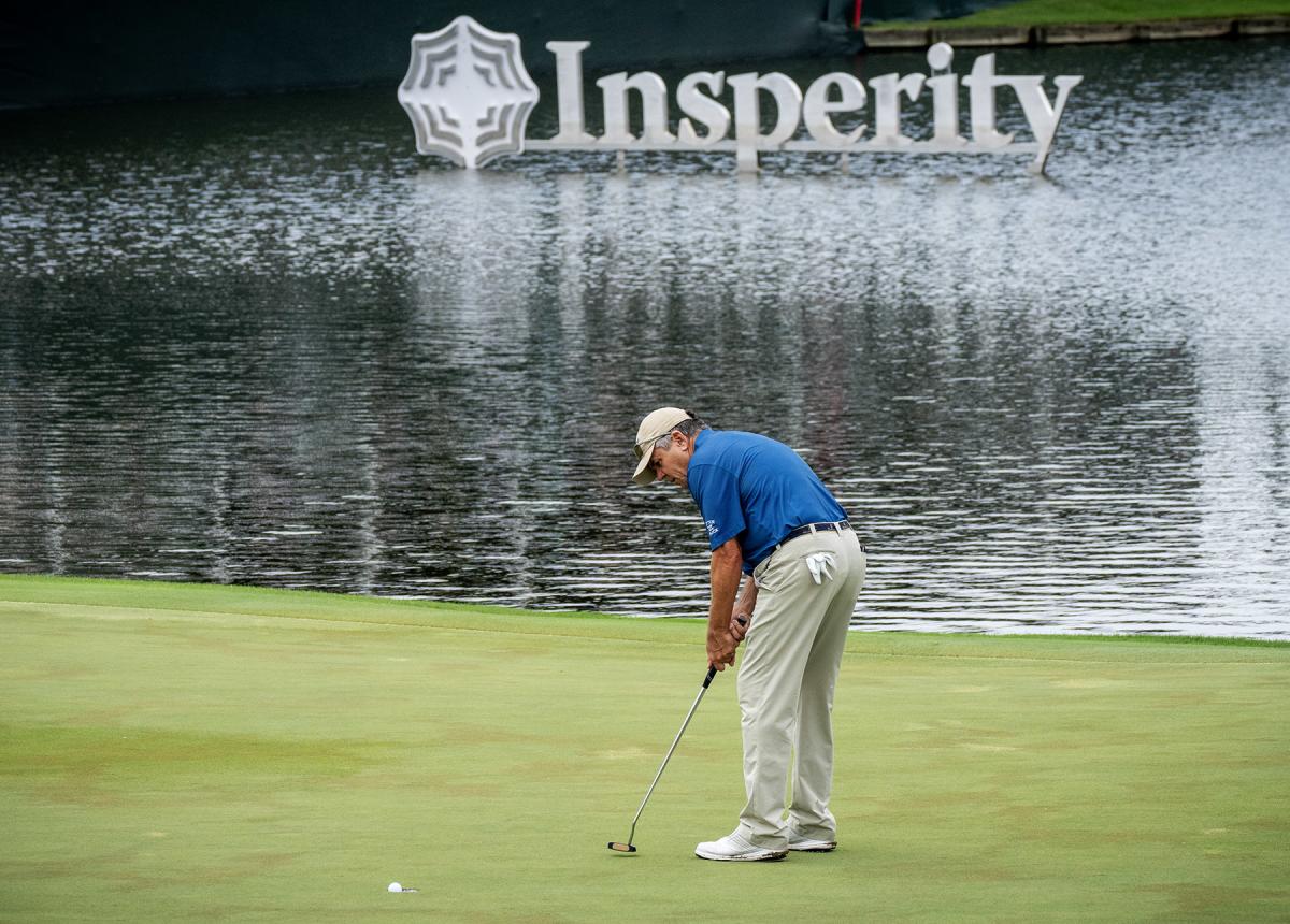 A white man, 50s-60s, putts a golf ball into the hole. He is wearing a blue sports polo, khaki slacks, white sneakers, and a khaki hat. A white glove is sticking out of his back pocket. Close behind him is a pond, and in the middle of the pond is a large white sign that reads "Insperity" with the competition's logo in front of the word.