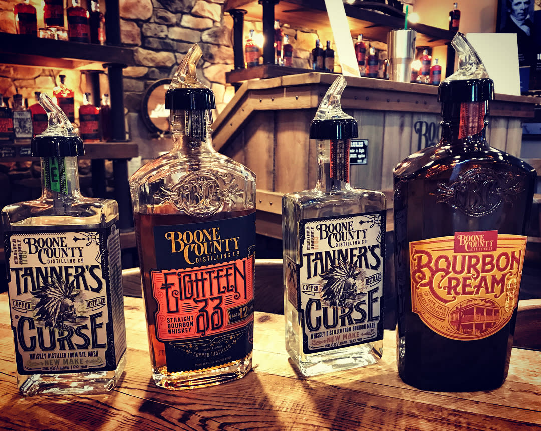 Two bottles of new make bourbon called Tanner's Curse, a brown bottle of Bourbon Cream and a bottle of 1833 bourbon on a wood table at Boone County Distilling Co.