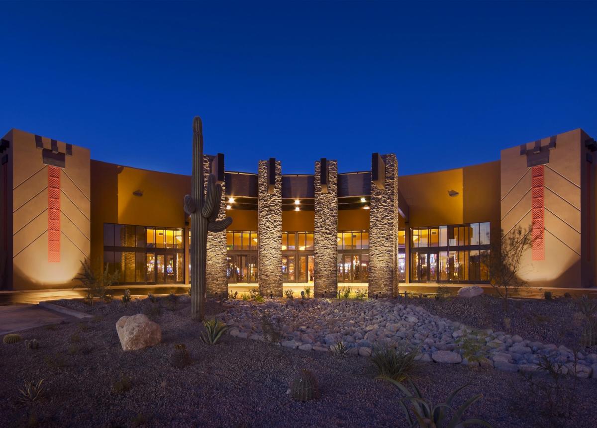 Exterior of Tucson's Desert Diamond Casino. A long, curved building with desert landscaping lit with golden glowing lights against dark night sky