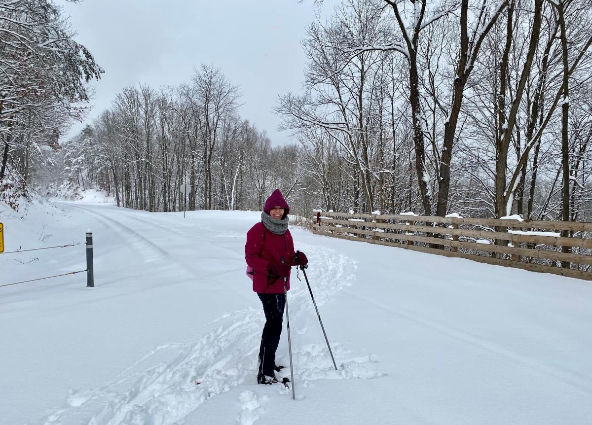 A woman is bundled up in winter gear and wearing corss country skis with poles in hand at a snow-covered trail head.