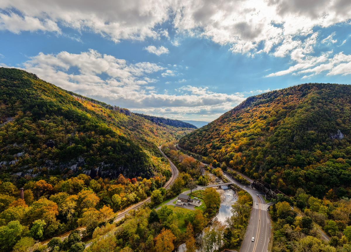 A cut in the mountain reveals a road, river and railroad, with bright blue skies and clouds and the pops of fall colors throughout the mountains.