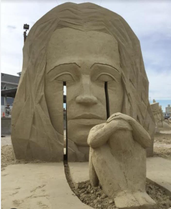 Sand sculpture of a woman's face. There is a cut where her tears run, down to the ground. The "puddle" of tears is actually a person curled up in the fetal position.