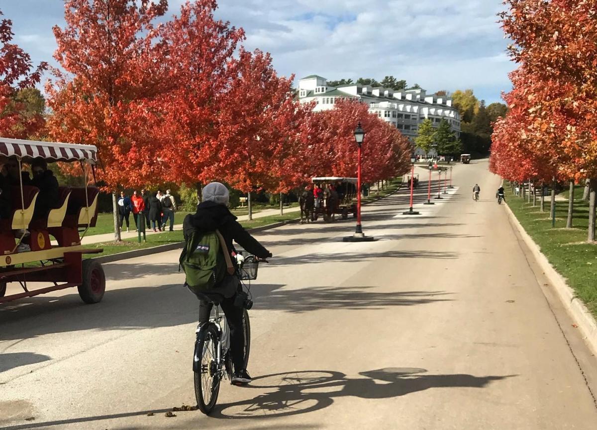 A woman wearing a backpack rides a bicycle up the road toward Mackinac Island’s Grand Hotel