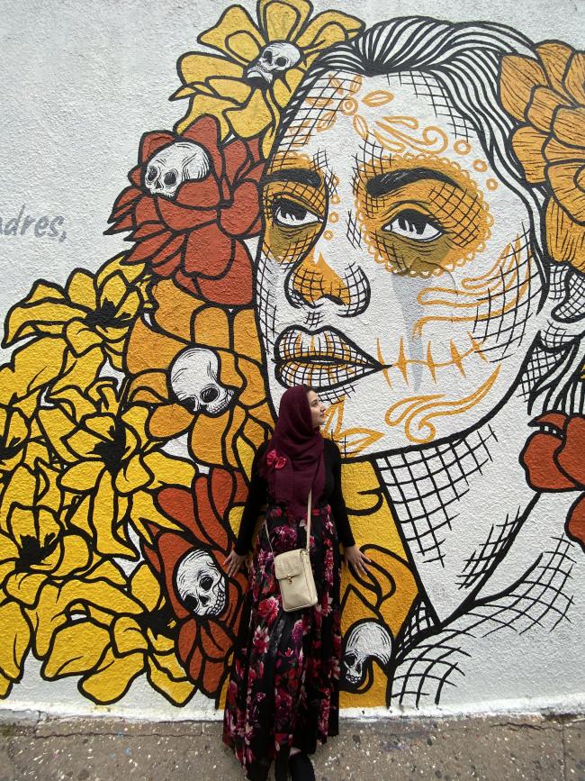 Miriam Ahmed stands in front of mural by Christin Apodaca at Mexic Arte Museum. She is wearing a floral, burgundy dress and hijab. The mural shows a crying woman wearing traditional Catrina makeup, surrounded by marigolds