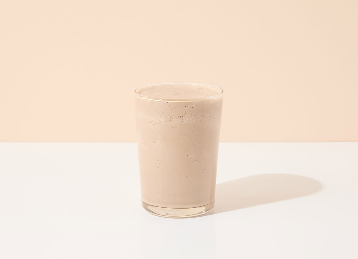 A simple glass of chocolate milkshake or protein smoothie set against a light beige background, with a smooth, creamy texture and a soft shadow on the surface.