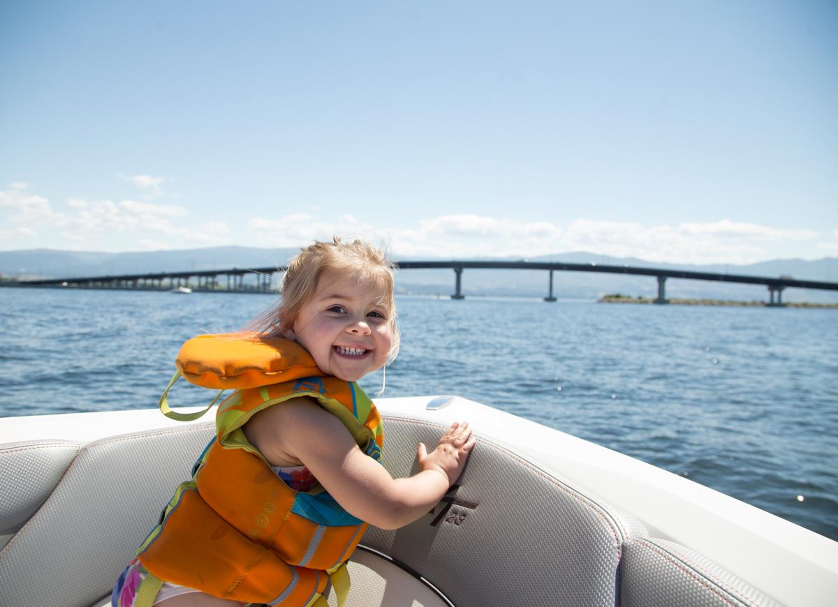 Young girl wearing a life jacket smiling while sitting on a boat, with the William R. Bennett Bridge and Okanagan Lake in the background on a sunny day