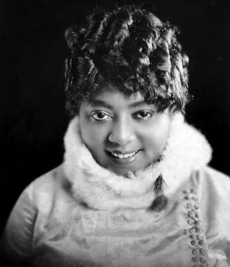 A woman faces the camera with a bright smile. She's wearing a light colored top with a fur collar.