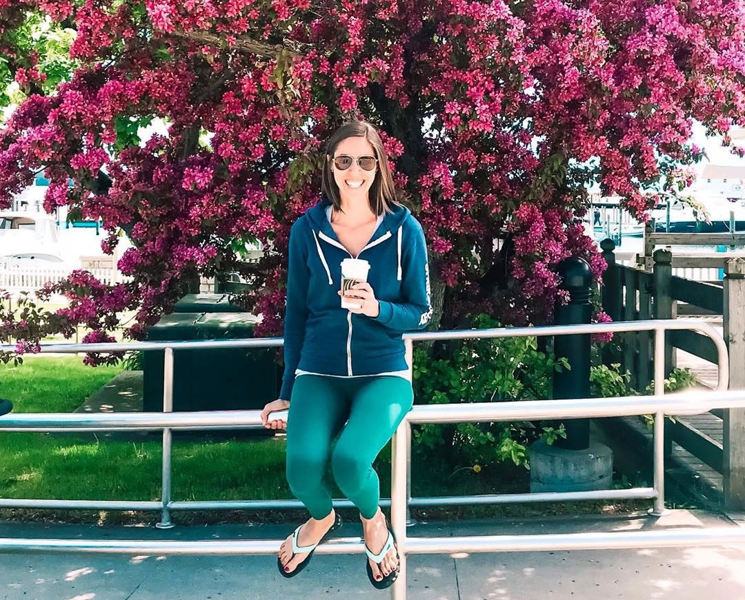 A woman holding a cup of coffee sits on a railing in front of a big, beautiful lilac bush in bloom