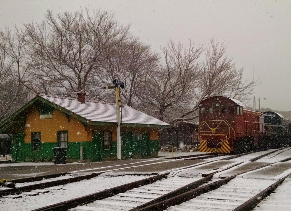 A small red diesel engine in a snowstorm next to a train station with a sign that says "Chase"