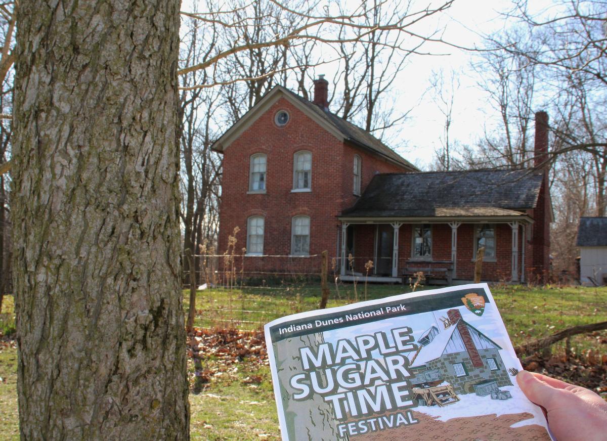 A hand holds a "Maple Sugar Time Festival" program in front of a historic building
