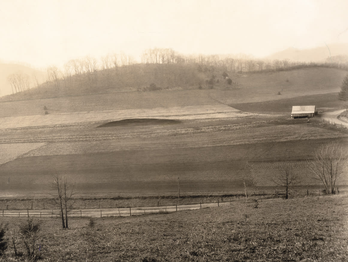A vintage, sepia-toned rural landscape photograph featuring a distinct, dark, flat-topped earthen mound situated in the center of plowed agricultural fields. A small wooden barn with a tin roof sits near a dirt road on the right side, with tree-lined hills rising in the background.