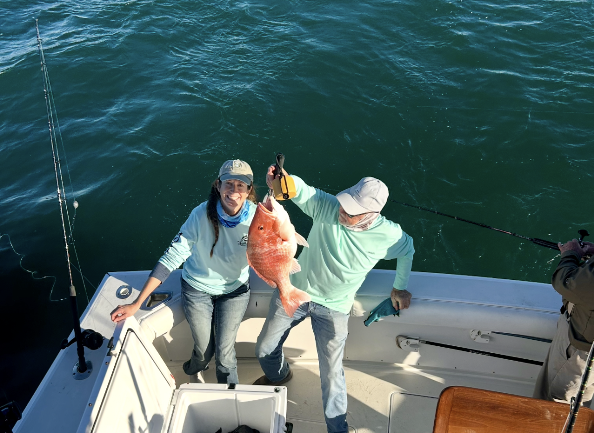 A woman and a man on the back of a boat holding a large red snapper fish.