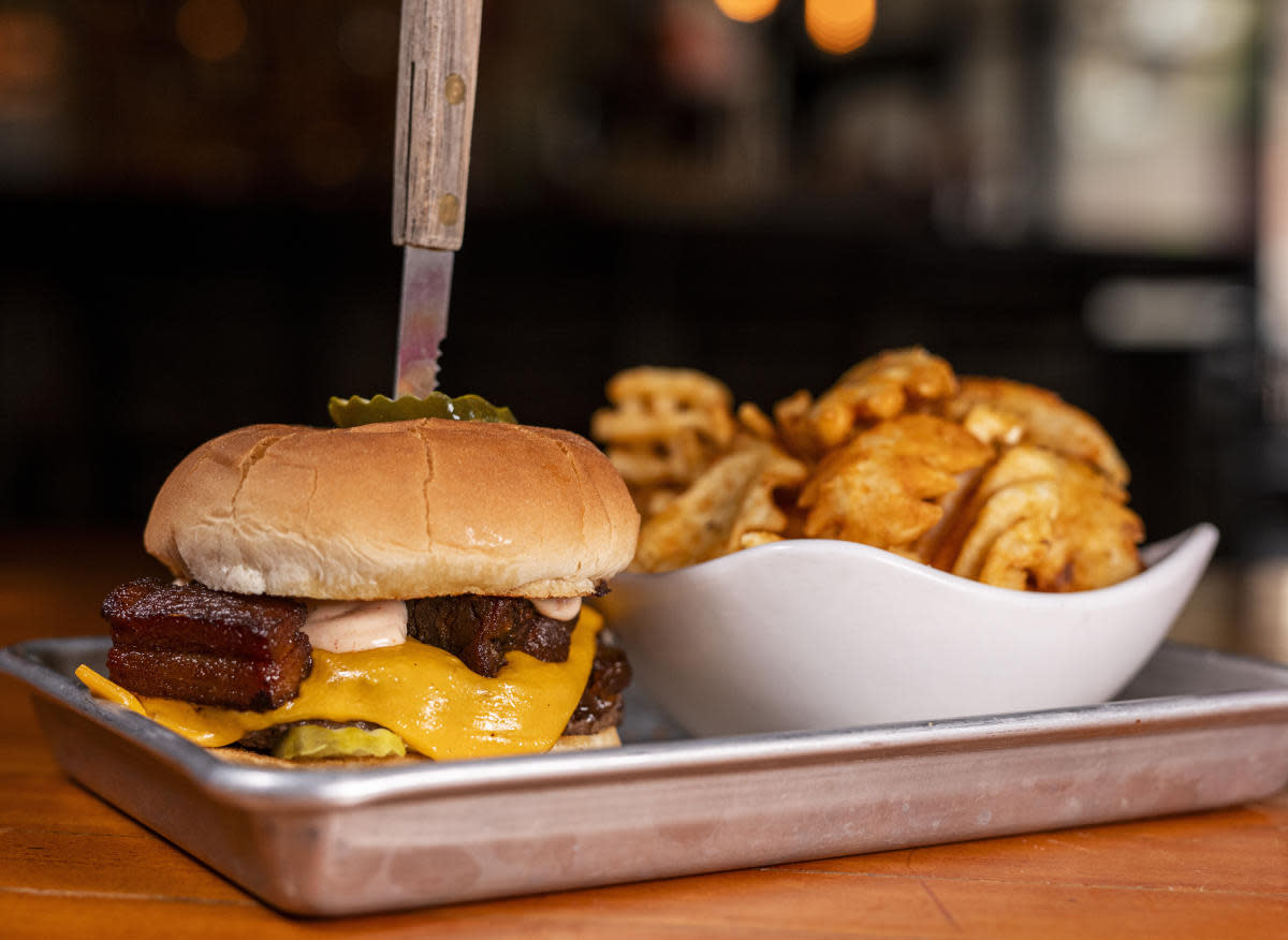 Cheeseburger with bacon, pickles, and sauce on a bun, served on a metal tray with a side of kettle chips in a white bowl.