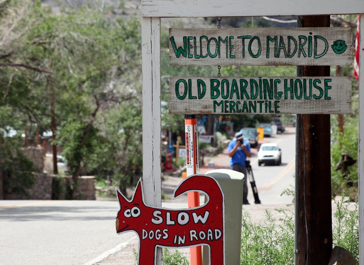 A sign on the side of a road reading Welcome to Madrid, Old Boarding House Mercantile, and a small sign below it depicts a dog and reads Slow Dog in Road