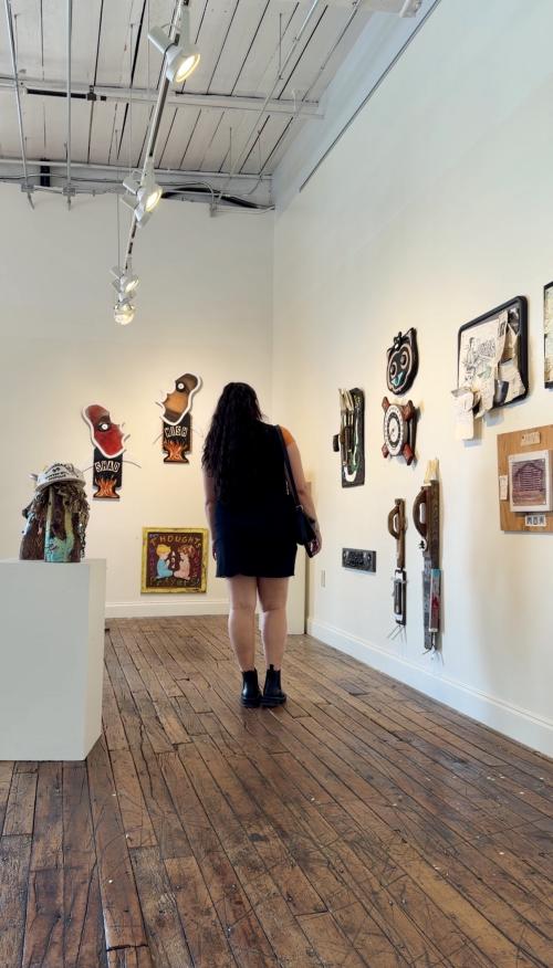 A visitor explores an art gallery at Lowe Mill ARTS & Entertainment, viewing colorful mixed-media artwork displayed on white walls beneath gallery lighting. The space features exposed wood floors and a variety of locally created pieces.