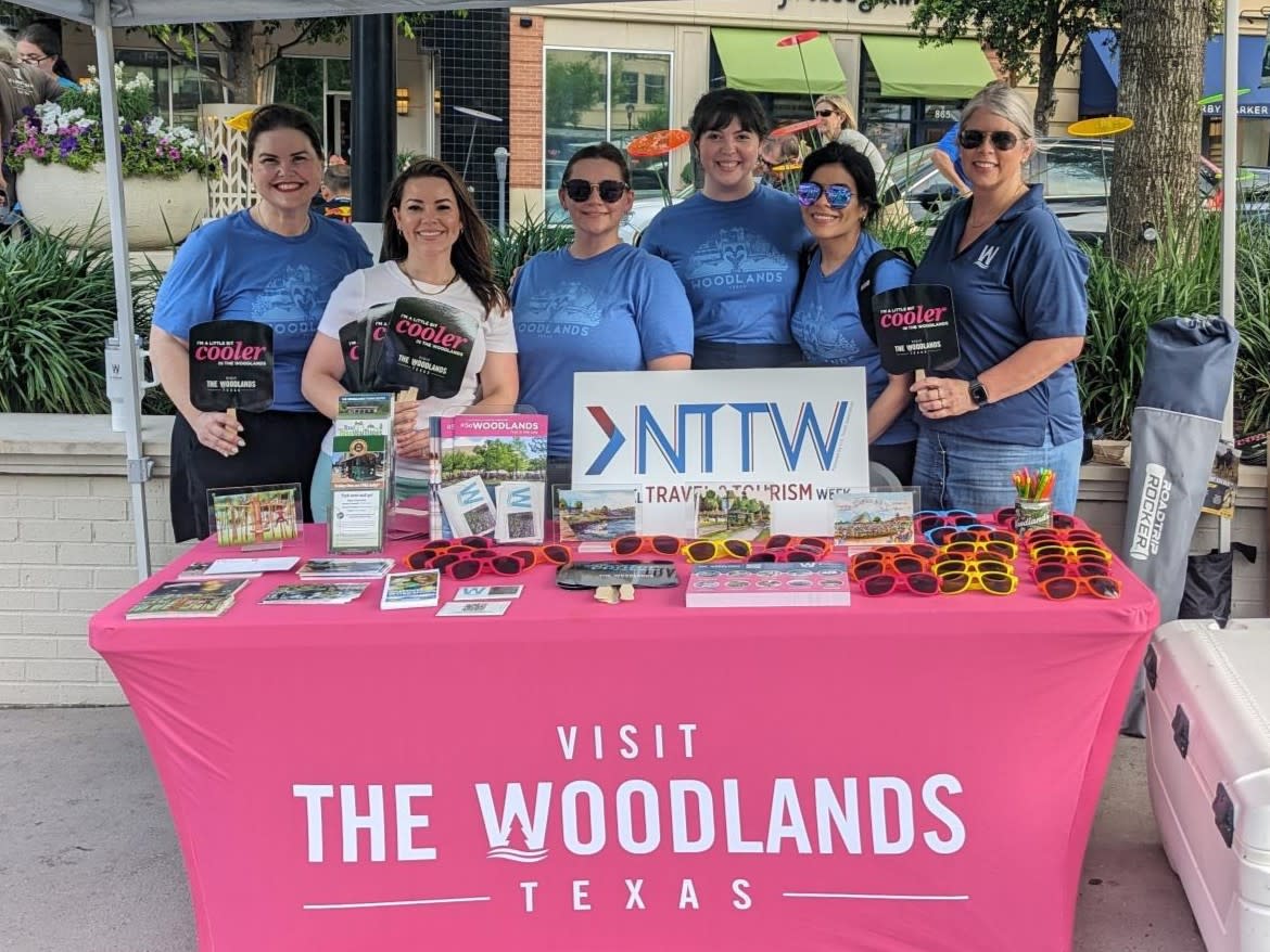 Six women stand behind a Visit The Woodlands booth at National Travel and Tourism Week (NTTW). Most of them are wearing matching blue T-shirts with an image of The Woodlands swan boats on them. Three are holding laminated paddles with wooden handles to hand out as fans for the event. The booth is covered in a hot pink cloth with "VISIT THE WOODLANDS TEXAS" in bold white text. On the table is a sign that says "NTTW" and features all kinds of giveaway swag (VTW brochures, maps of The Woodlands, sunglasses, postcards, etc.).