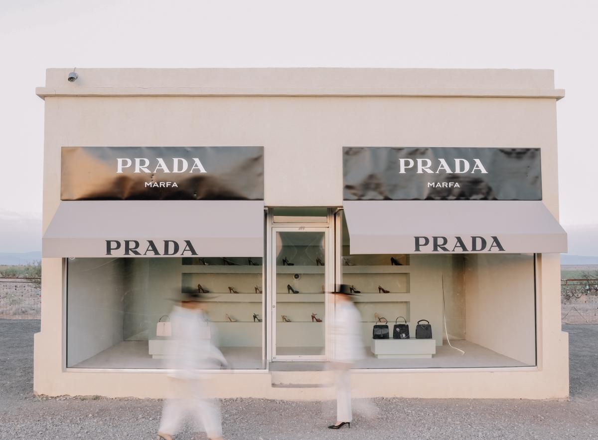 A beige, minimalist building labeled “Prada Marfa” stands alone in the desert, styled like a high-fashion boutique with large glass windows displaying luxury shoes and handbags. Two blurred figures in white walk past, adding motion to the still West Texas landscape.