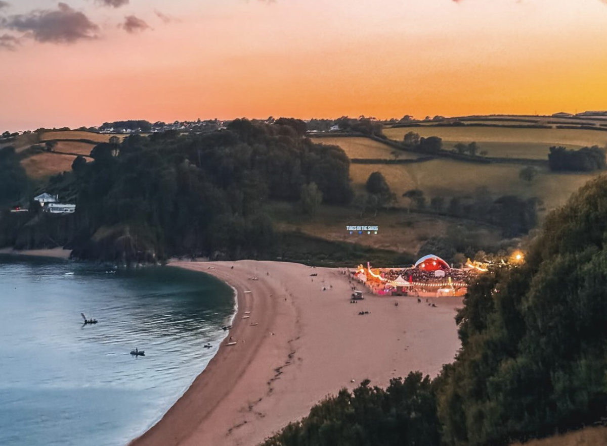 Aerial view of a vibrant beach festival at sunset with glowing lights and a red dome stage. Calm sea, scattered visitors, and lush, rolling hills.