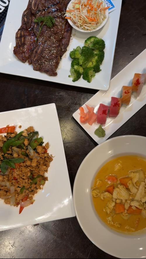 An overhead view of a variety of dishes at Phuket Thai Restaurant in Huntsville, Alabama, including a grilled steak with broccoli and coleslaw, sushi, spicy basil chicken, and a bowl of yellow curry with chicken and vegetables.