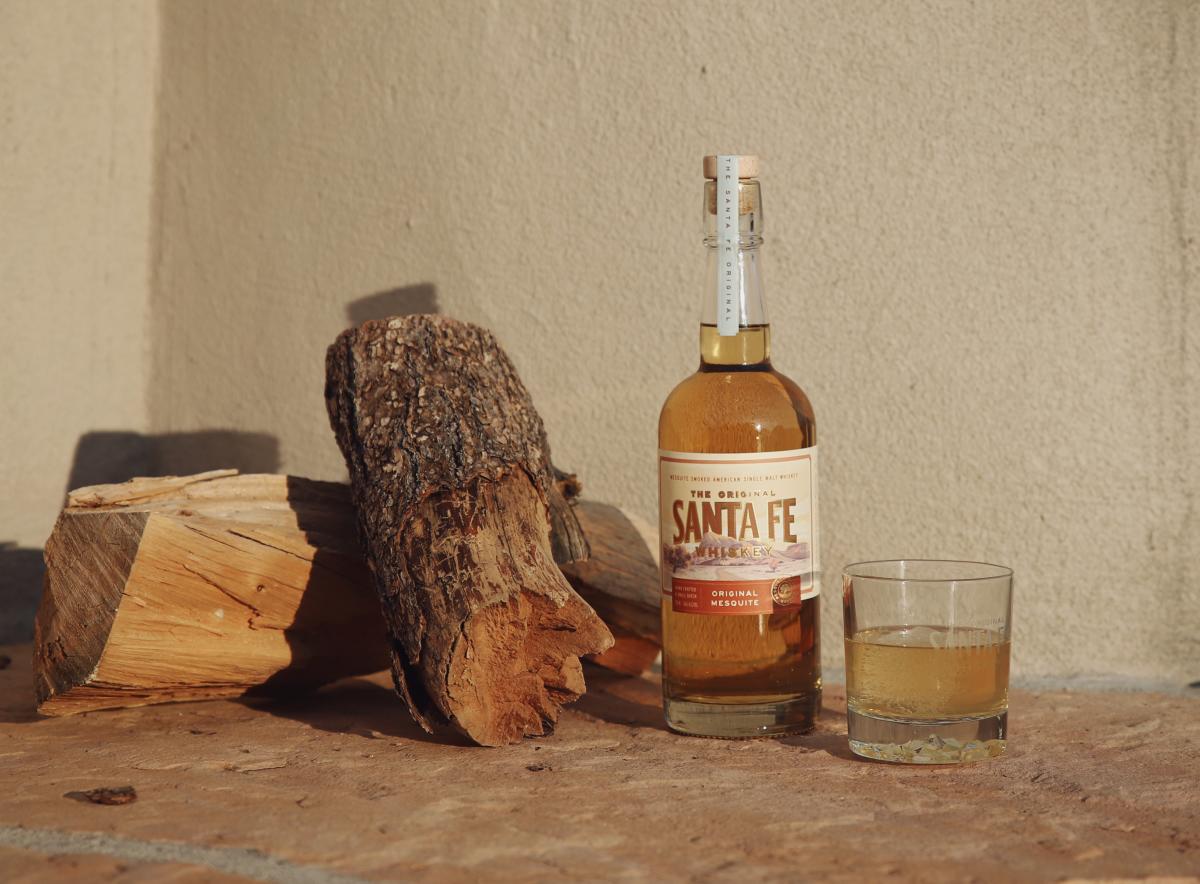 A bottle of Santa Fe whiskey next to a glass half-filled with a golden liquid, set alongside some chopped firewood against a sunlit wall.