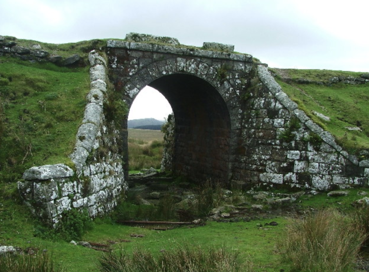Ancient stone arch bridge overgrown with moss in a lush green landscape. The scene feels serene and timeless, set under a cloudy sky.