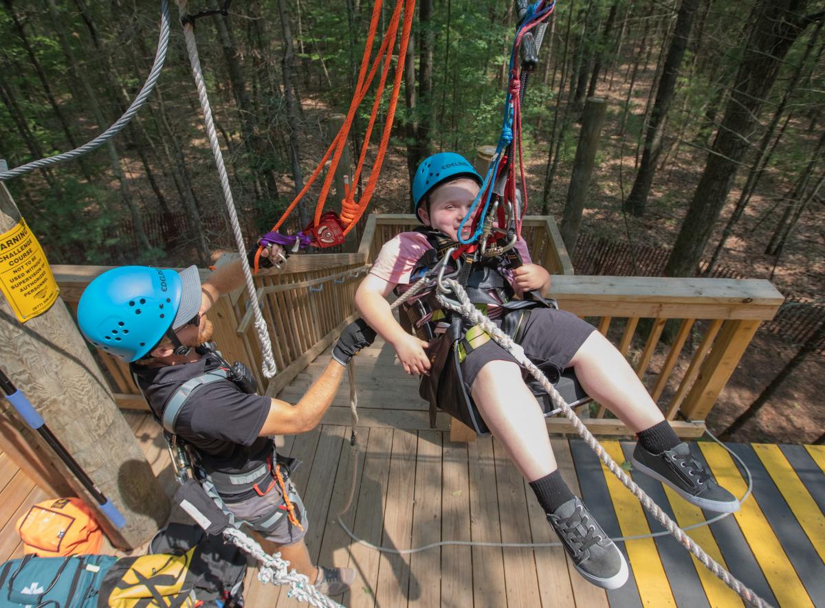 Two individuals wearing helmets and harnesses prepare for a zip-lining adventure in a forested area.