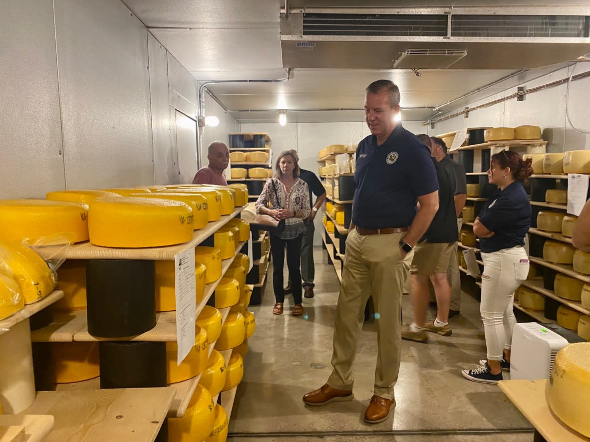 NYS Assemblyman standing in cheese room at Muranda Cheese surrounded by solid wheels of cheddar
