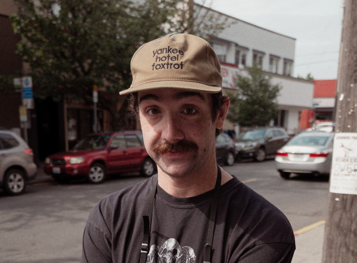 John Malone, wearing a tan Yankee Hotel Foxtrot hat and dark shirt, stands outside on a Milwaukee street.