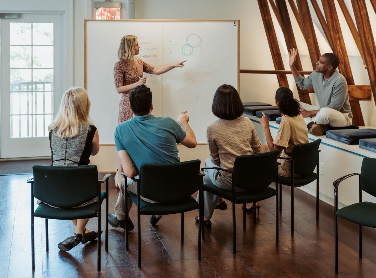 A group of people are meeting in a room with a dry erase board. One person is raising his hand to ask a question.