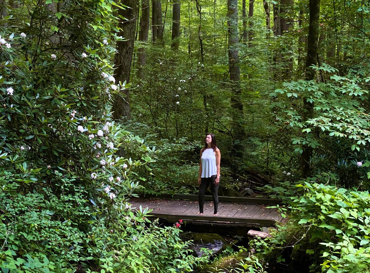 A woman stands peacefully with her eyes closed on a wooden footbridge over a small stream, surrounded by dense green forest foliage and blooming white rhododendrons.