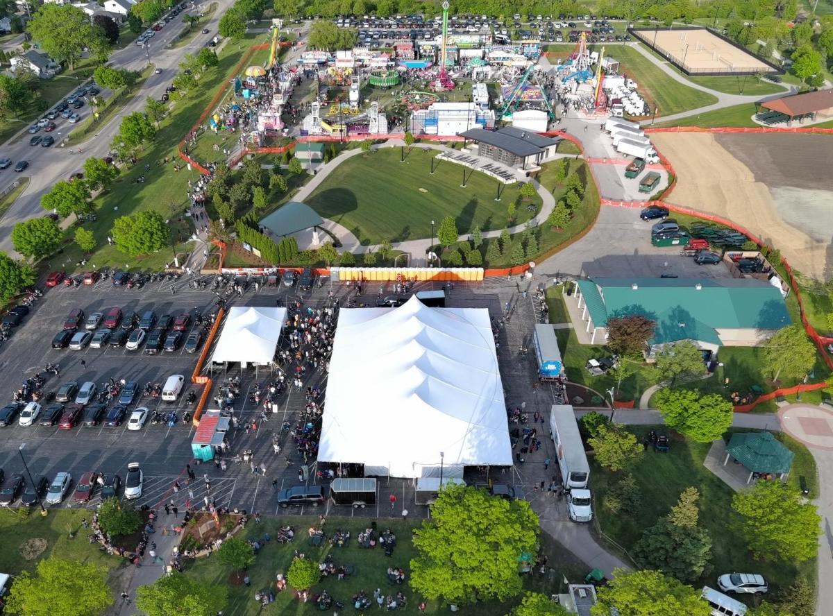 Aerial view of a large outdoor festival with a carnival setup, including rides, food stands, and game booths arranged in a park. A large white event tent is filled with people in the foreground, with lines of parked cars and crowds gathering around it. Pathways, green lawns, and nearby buildings surround the event, with busy streets and additional parking visible along the edges.