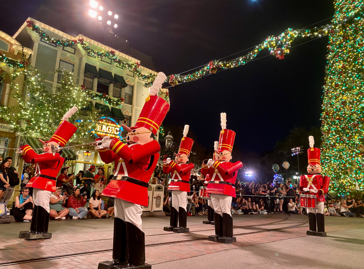 Image of the Disneyland Resort holiday parade walking down Main Street U.S.A. In the background, a large Christmas tree decorated with green, yellow, and red lights can be seen. There are also store fronts in the back left-hand corner with guests standing in front of the stores watching the parade.