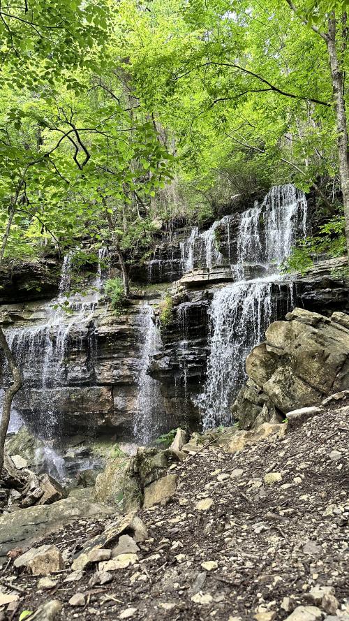Bethel Spring Falls cascades over layered rock formations surrounded by lush green trees. The scenic waterfall flows into a rocky creek below, creating a peaceful setting along the forest trail.
