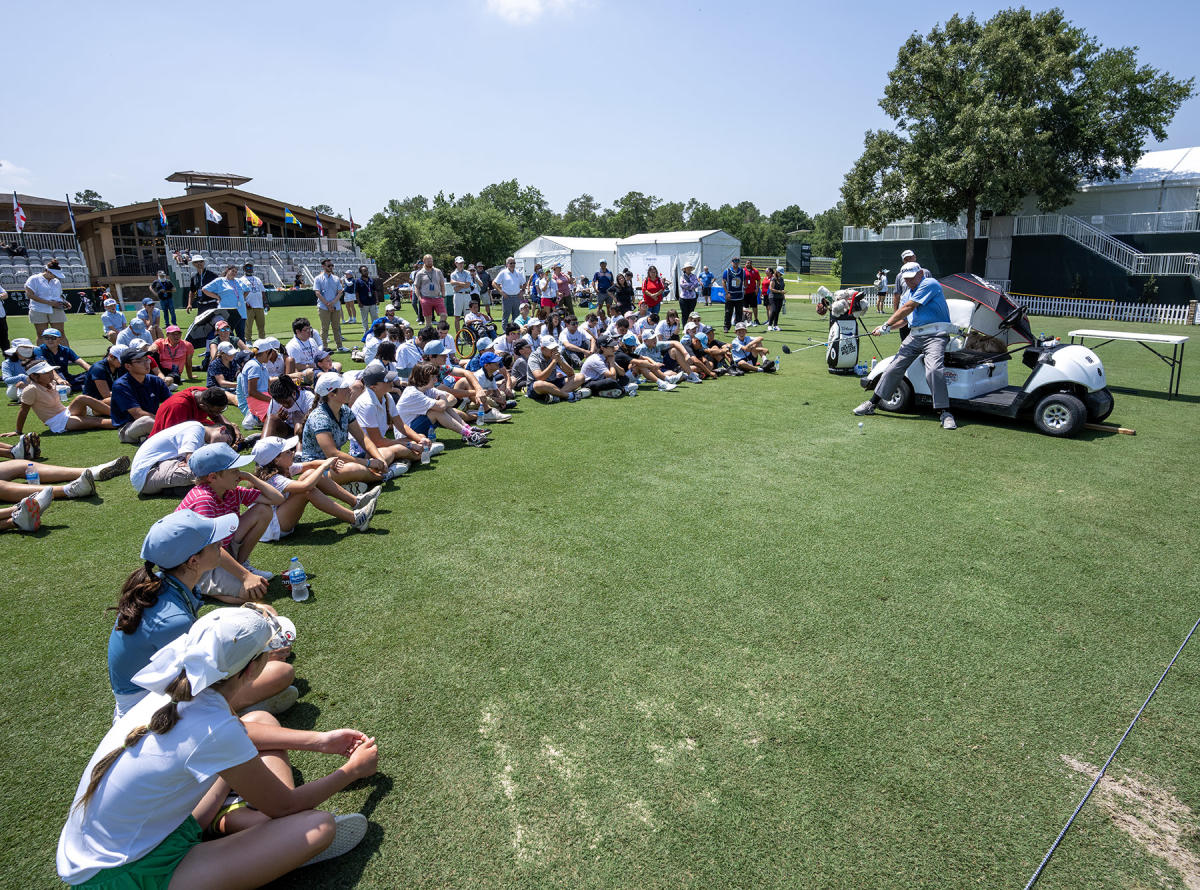 A group of 40+ children and teens sit in a semicircle around a pro golfer demonstrating his swing.