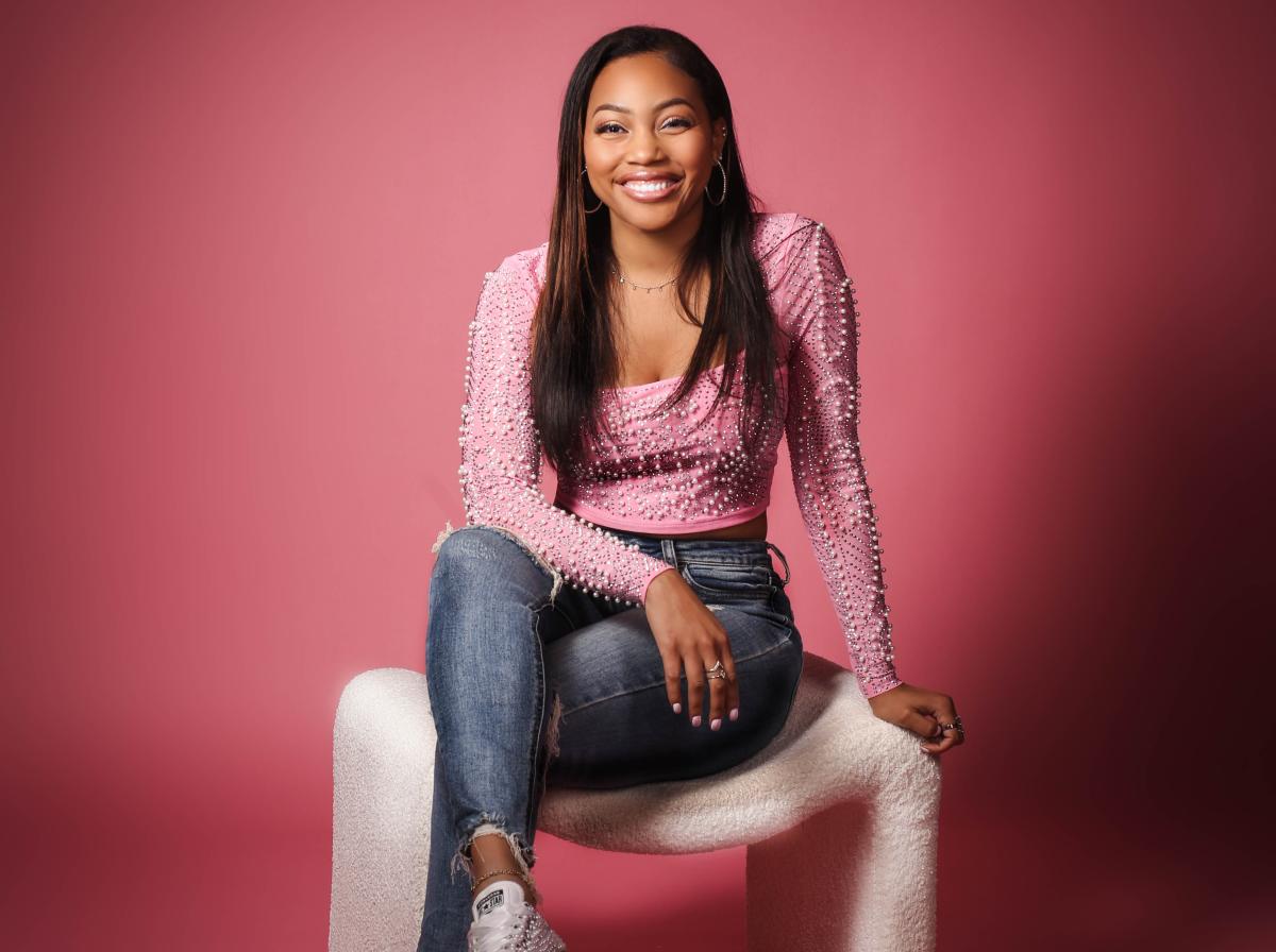 Social media influencer Jasmine Jennings sits on a white bench in front of a pink background, wearing a pink shirt with pearls, jeans and sparkly tennis shoes.
