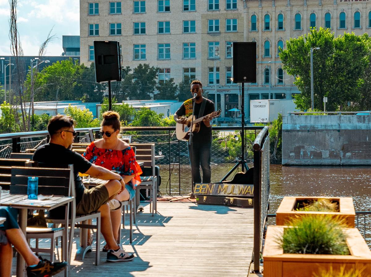 A musician performs with an acoustic guitar on a riverside patio while seated guests enjoy drinks at nearby tables. Two speakers frame the performance area, and a guitar case with the performer’s name sits at the front. A river, trees and historic downtown buildings create the backdrop on a sunny afternoon.