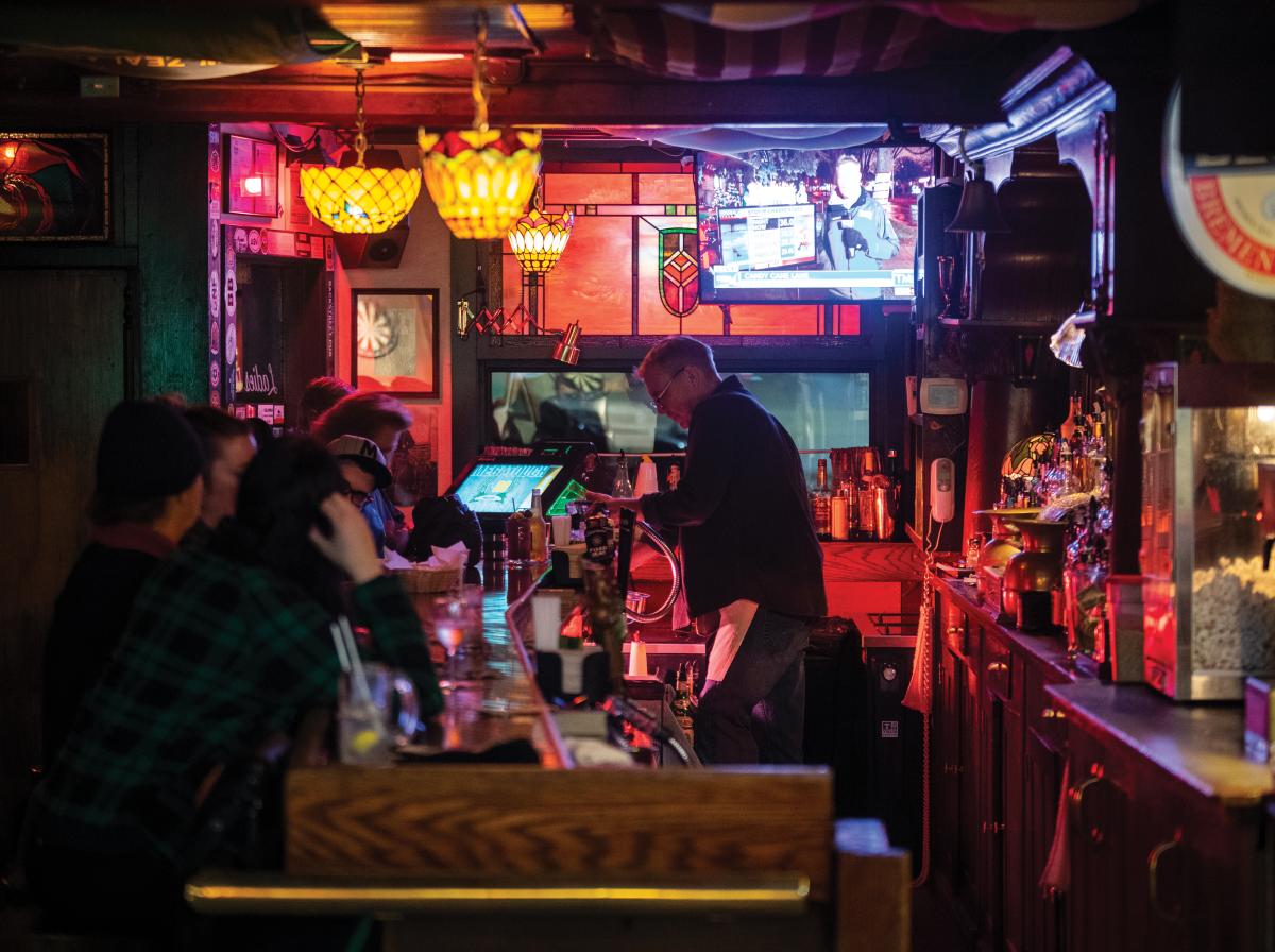 Inside Wolski’s Tavern, a cozy Milwaukee bar with warm red and yellow lighting. Several patrons sit at the wooden bar counter while the bartender serves drinks. Stained glass lamps hang overhead, bottles line the back wall, and a TV shows a news broadcast, creating a classic neighborhood pub atmosphere.