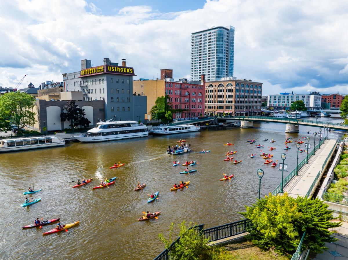 Dozens of kayakers paddle along the Milwaukee River past downtown buildings, boats and the RiverWalk on a sunny day.