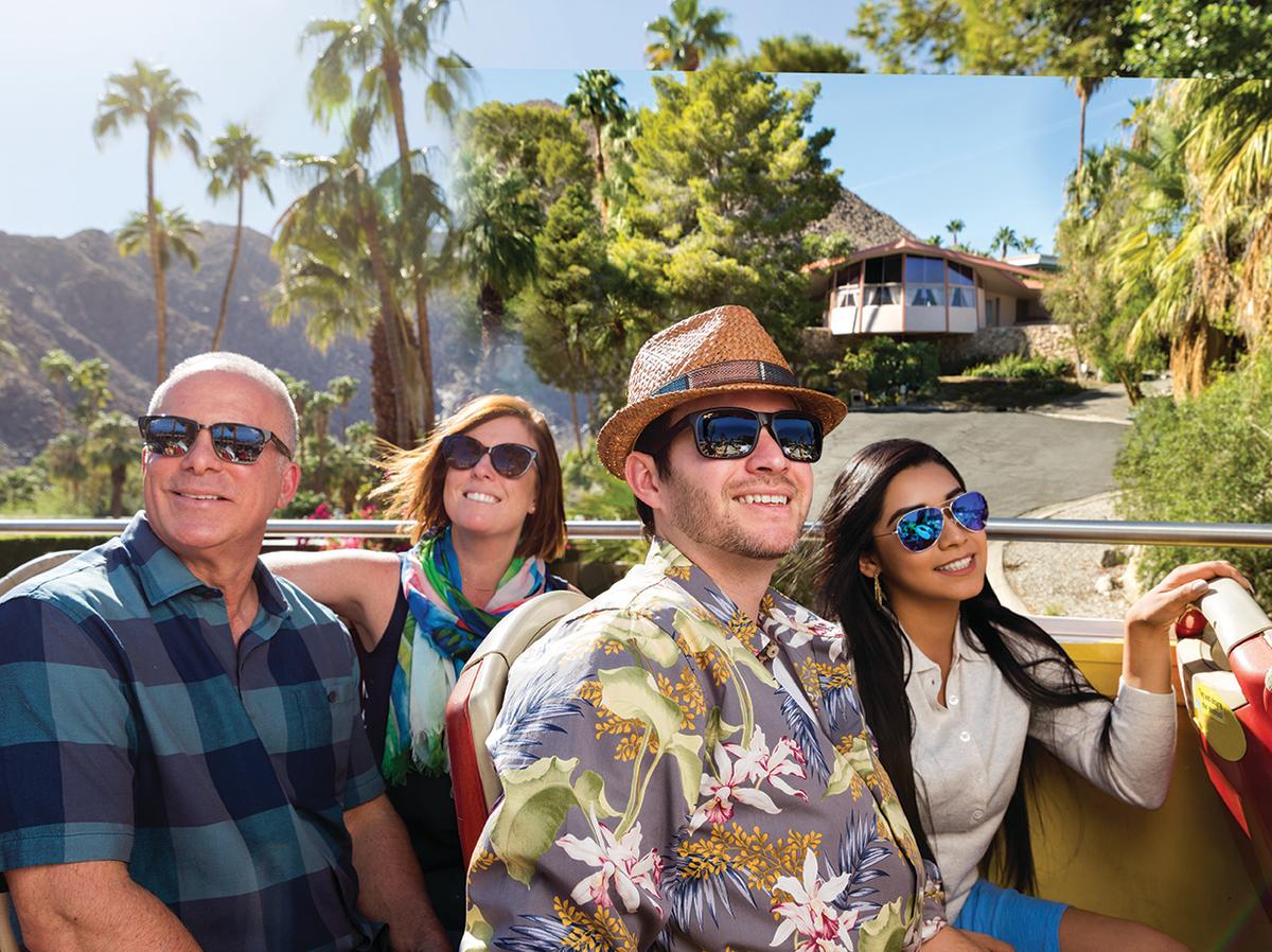 Four people wearing sunglasses are smiling and enjoying a sunny day on an open-top tour bus. Palm trees and mountains are in the background with a mid-century modern house nestled among the trees.